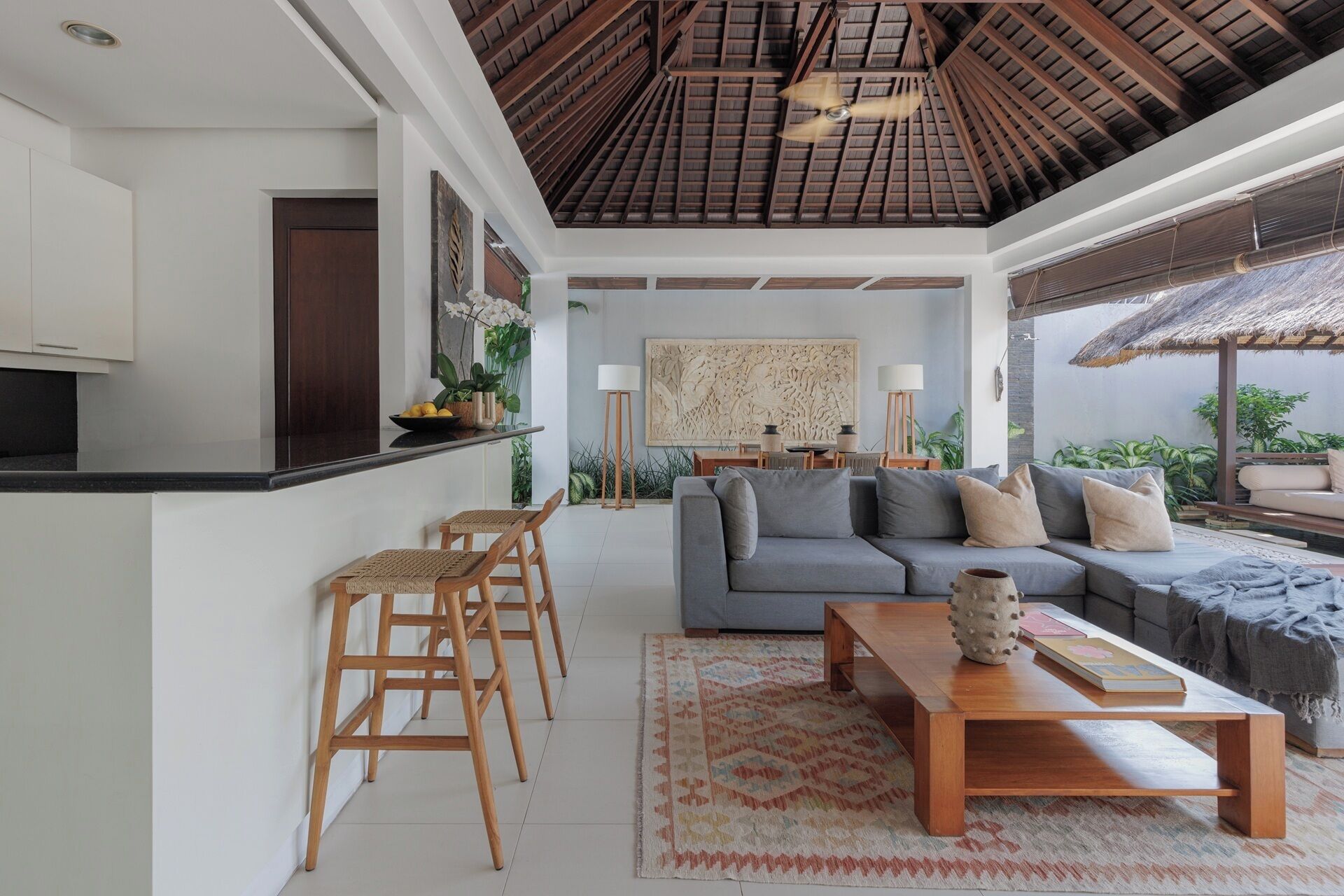 A modern living space with a kitchen, a grey sofa, wooden chairs, a coffee table, and a dining area.