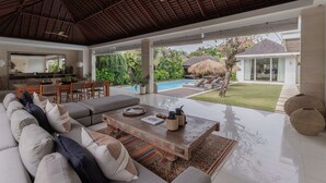 A spacious living area with a large sofa, a wooden coffee table, and a view of a swimming pool and garden. - Chandra Bali Villas (Seminyak)