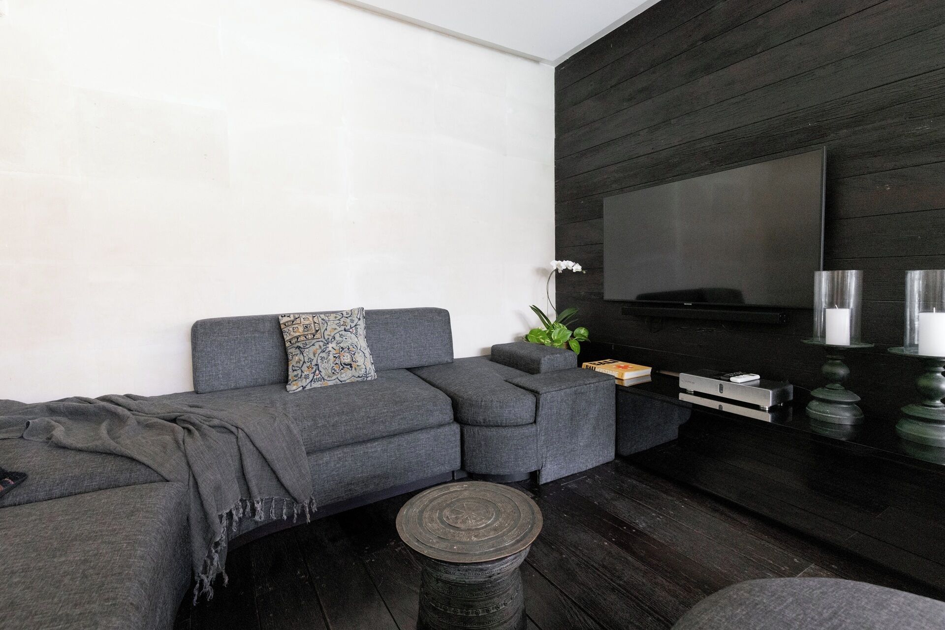 A modern living room with a grey sofa, a small round table, a flat-screen TV mounted on a dark wall, and a potted plant.