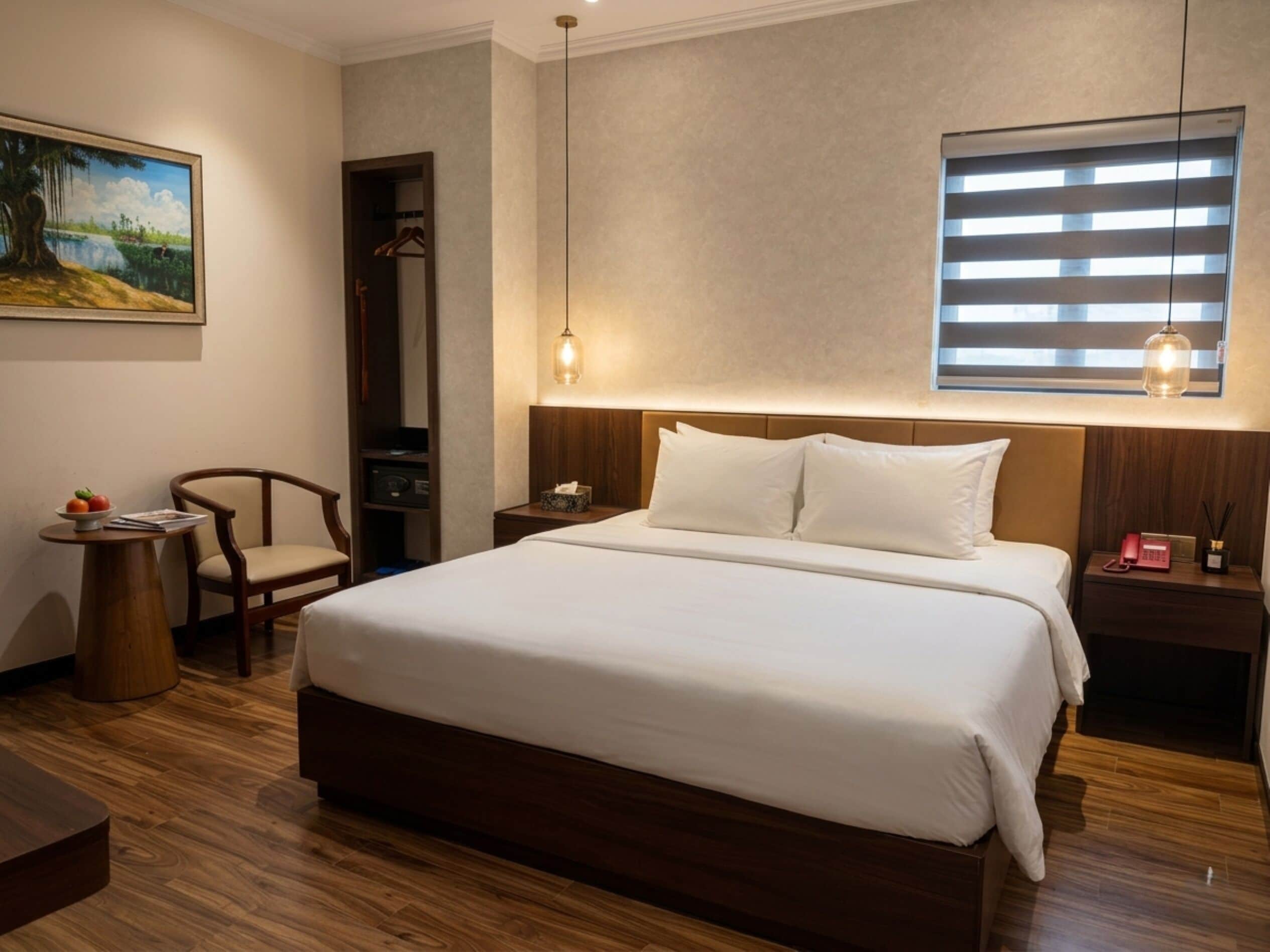 Quarto de hotel com uma cama grande, piso de madeira, um quadro na parede, uma cadeira, uma pequena mesa com frutas e uma janela com persianas.