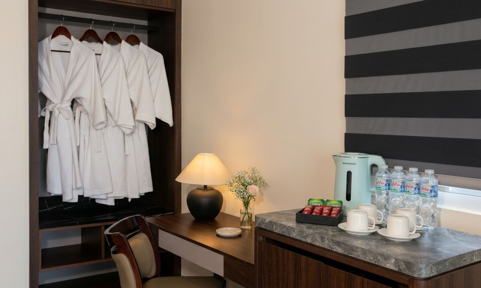 A hotel room with a robe rack, a lamp, a coffee station, and a window with blinds.
