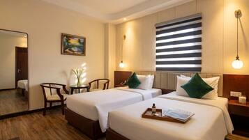 A hotel room with two beds, a desk, a chair, a painting, and a window with blinds.