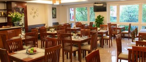A dining area with multiple wooden tables and chairs, a kitchen area with a counter, and large windows allowing natural light.