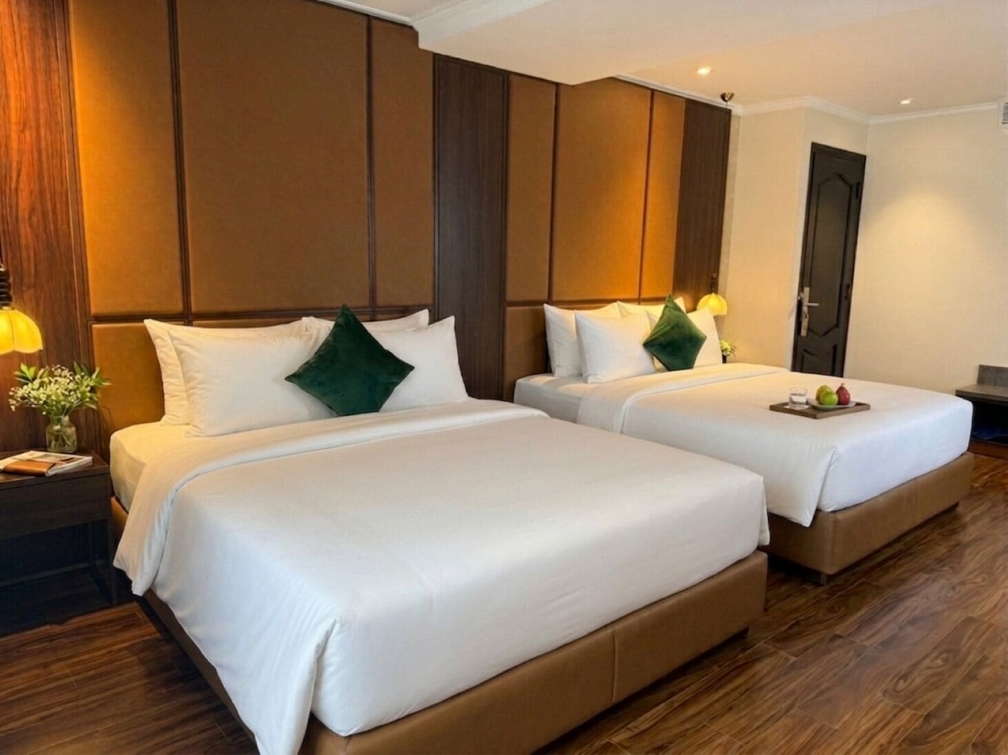 Habitación de hotel con dos camas, cabeceros de madera, una mesita de noche con lámpara y una bandeja de frutas sobre la cama.