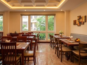 A dining area with wooden tables and chairs, large windows with a view of greenery, and artwork on the walls.