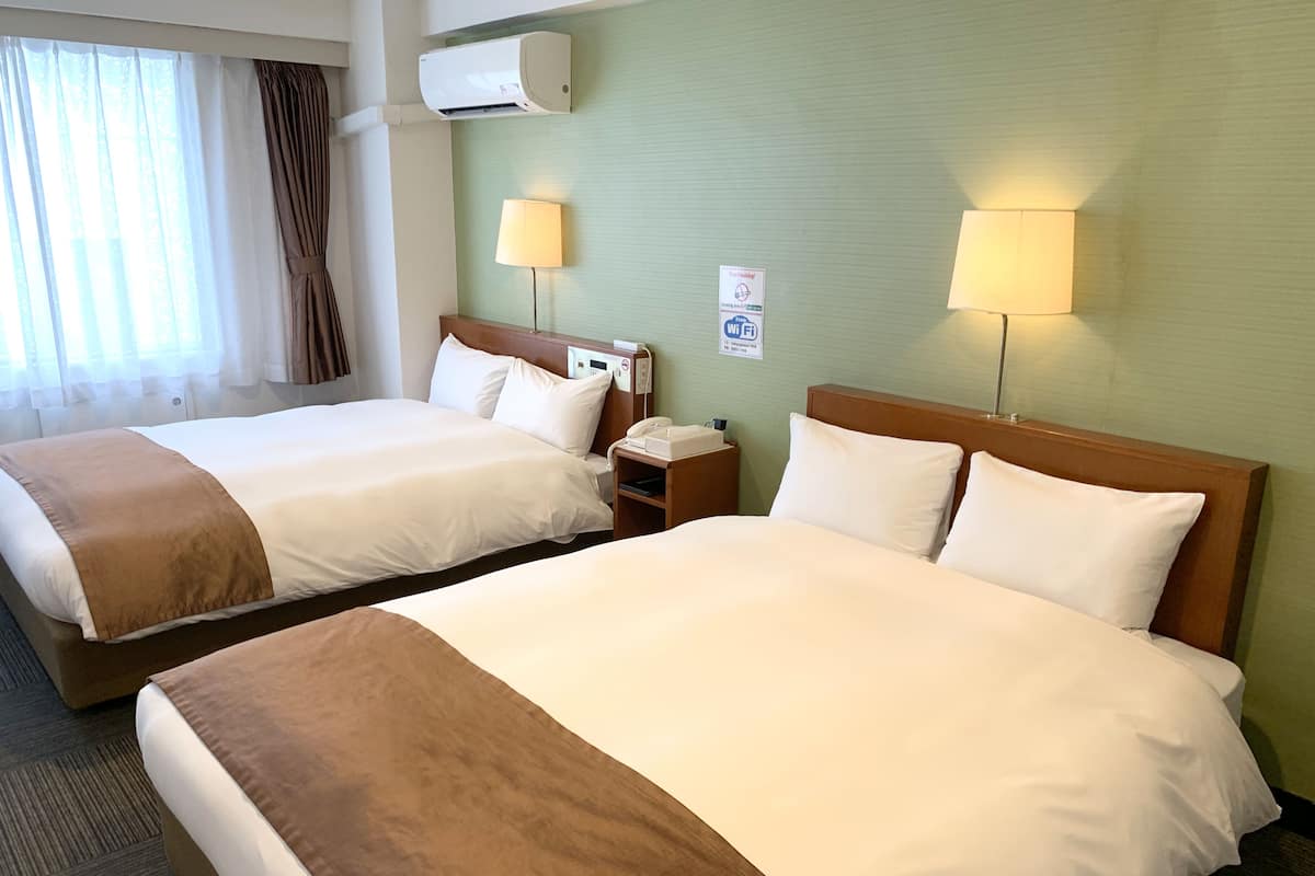Habitación de hotel con dos camas, una mesita de noche con lámpara, teléfono y aire acondicionado.