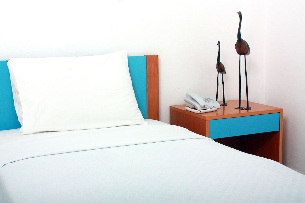 Una cama bien hecha con un almohadón blanco y una mesita de noche de madera con un cajón azul, un teléfono blanco y dos esculturas decorativas de pájaros.
