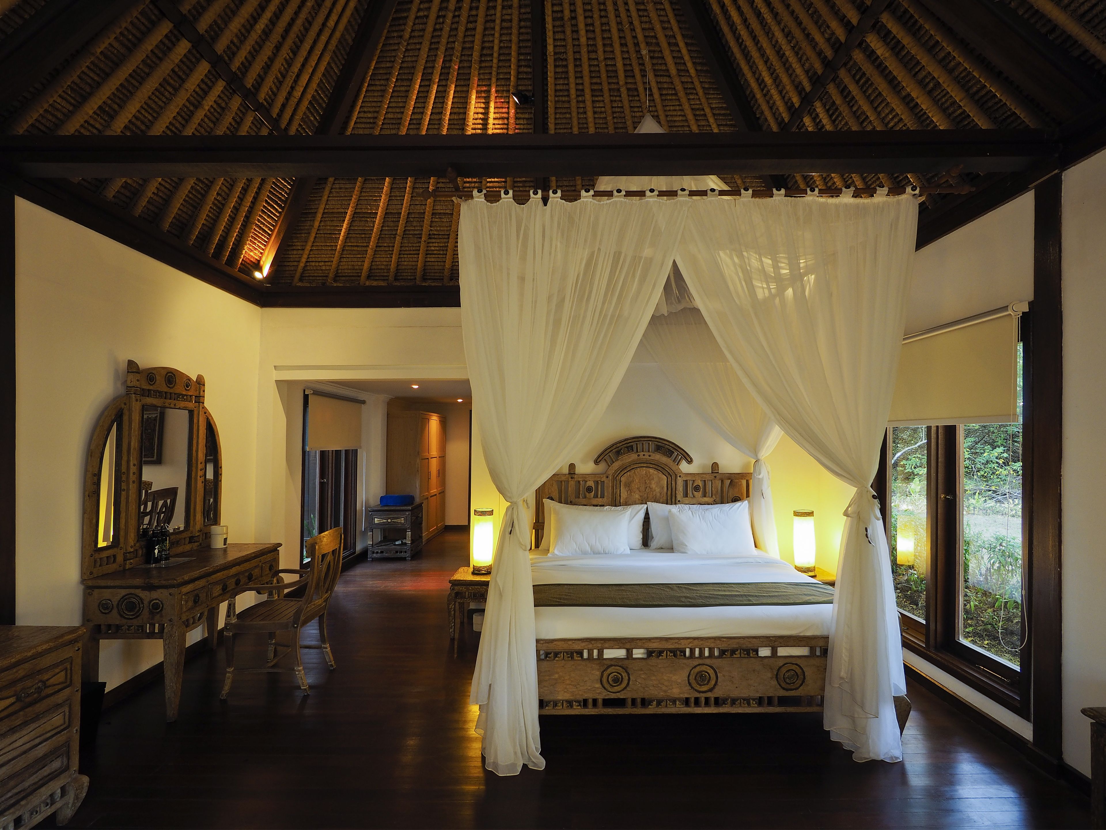 Un dormitorio amplio con una cama grande, cabecera de madera y dosel. Hay un aparador de madera, un espejo y una ventana con vistas a zonas verdes.