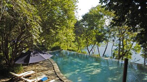 Piscina com espreguiçadeiras e guarda-sol, rodeada por árvores e um corpo de água.