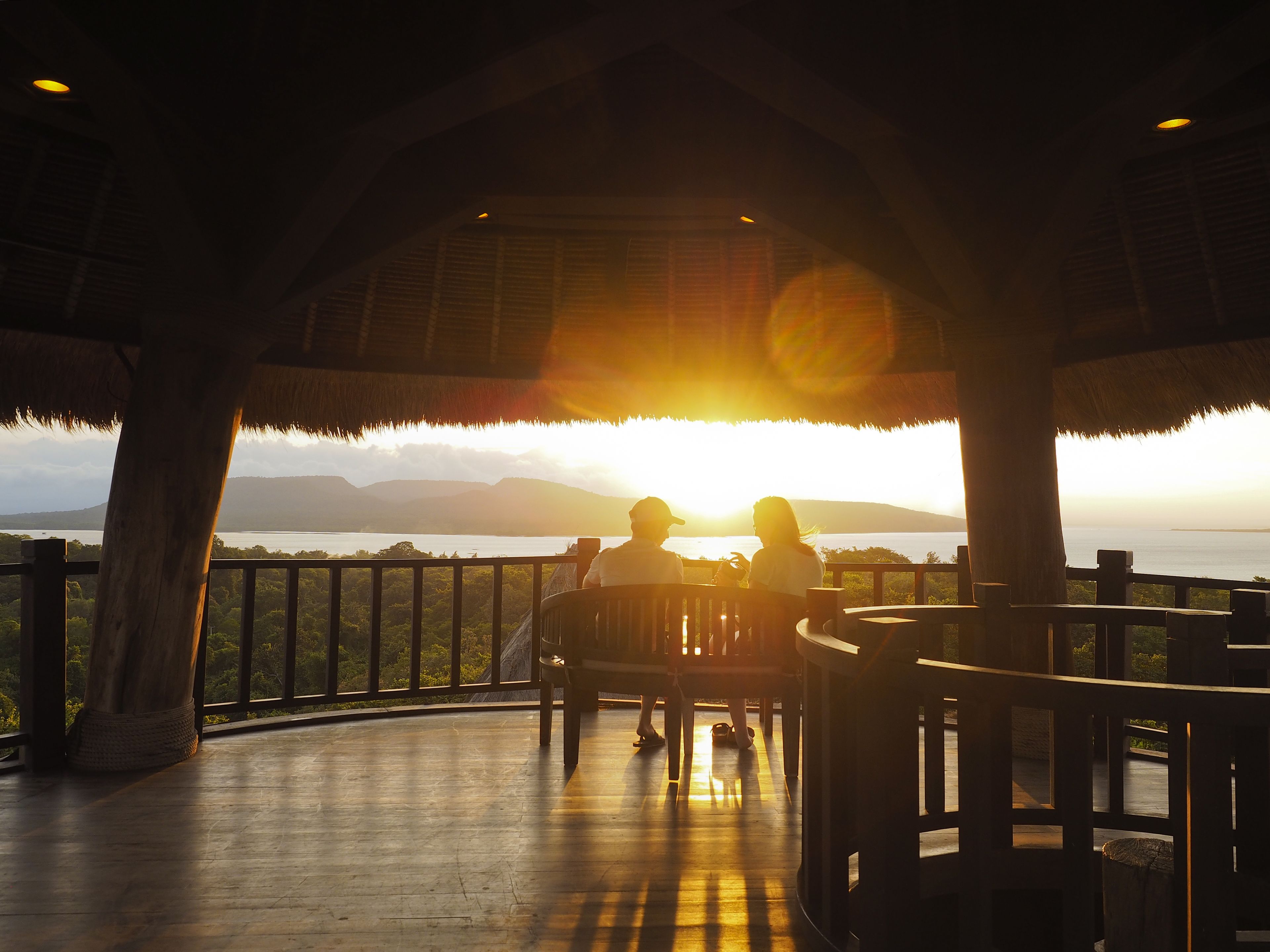 A couple sitting on a wooden deck with a view of the sunset over a body of water and mountains.