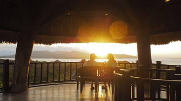 A couple sitting on a wooden deck with a view of the sunset over a body of water and mountains.
