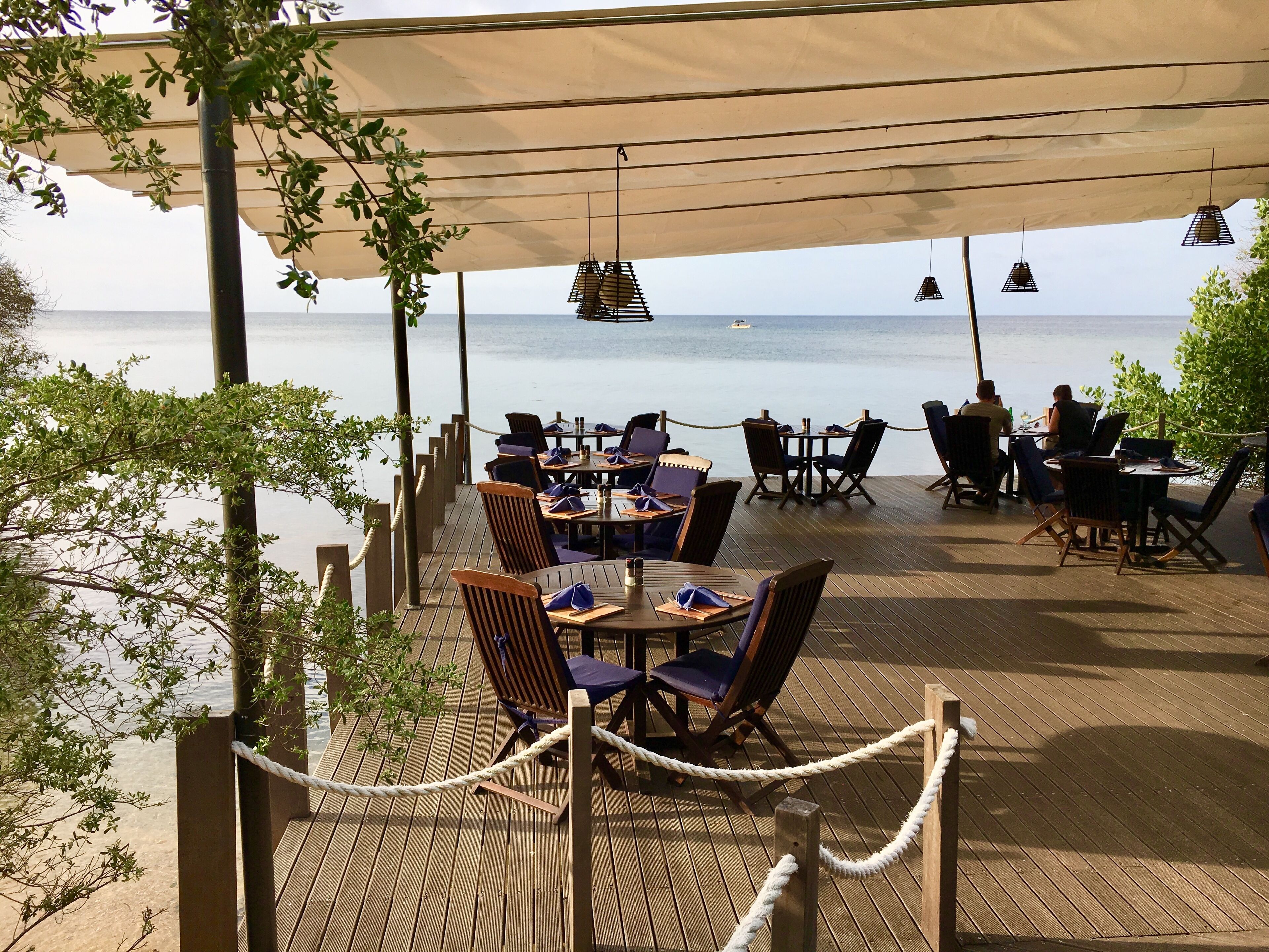 Un área de comedor al aire libre con mesas y sillas, vista al mar y un toldo que da sombra.
