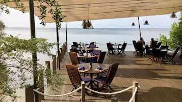 Un área de comedor al aire libre con mesas y sillas, vista al mar y un toldo que da sombra.