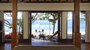 Quarto com uma cama grande, pilares de madeira e vista para uma praia com cadeiras e um guarda-sol.