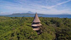Uma construção tradicional com telhado de palha, rodeada por uma floresta verde densa, com vista para um corpo d'água e montanhas distantes.