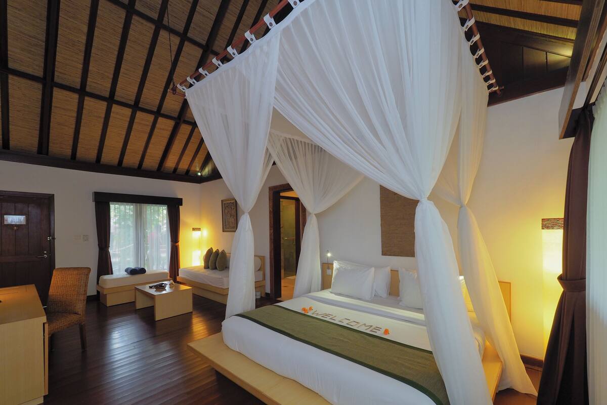 A bedroom with a canopy bed, a sitting area with a chair and table, and a wooden floor.