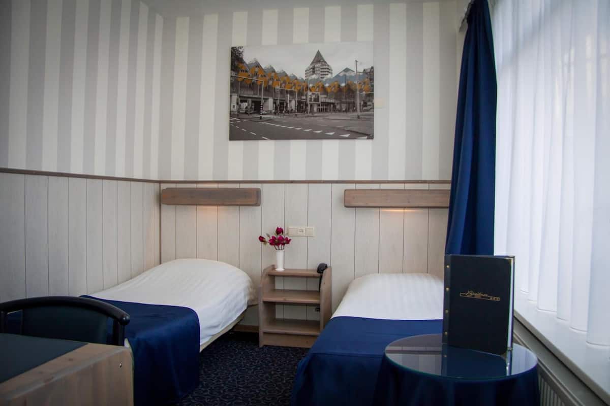 A hotel room with two single beds, a small table, a chair, a nightstand, a vase with flowers, a framed picture on the wall, and a window with curtains.