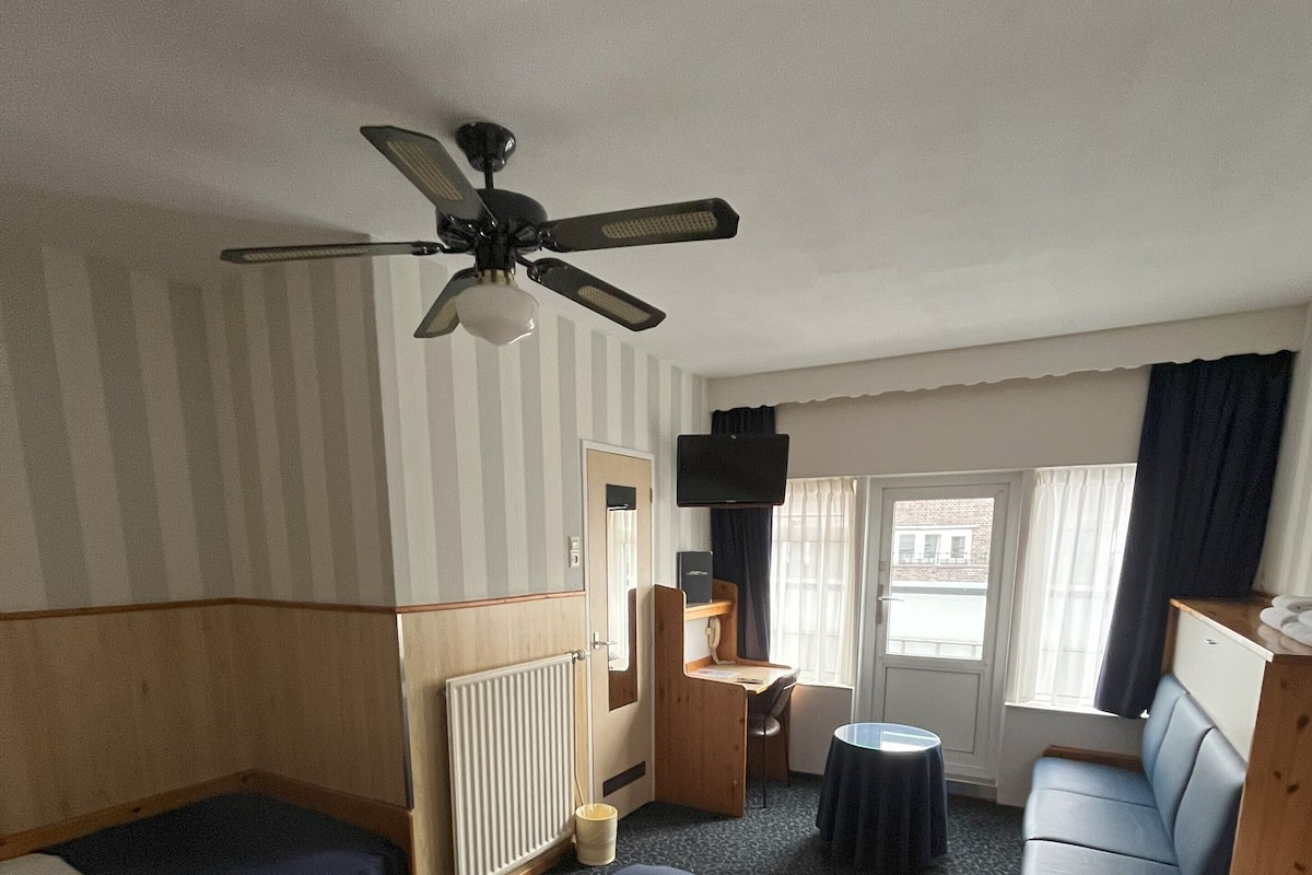 A hotel room with a large bed, two single beds, a desk, a chair, a sofa, a ceiling fan, and a window with curtains.