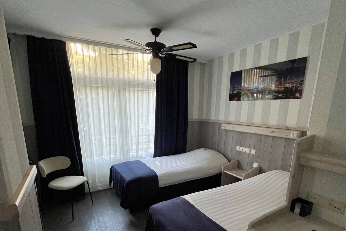 A hotel room with two single beds, a desk with a chair, a ceiling fan, and a window with curtains.