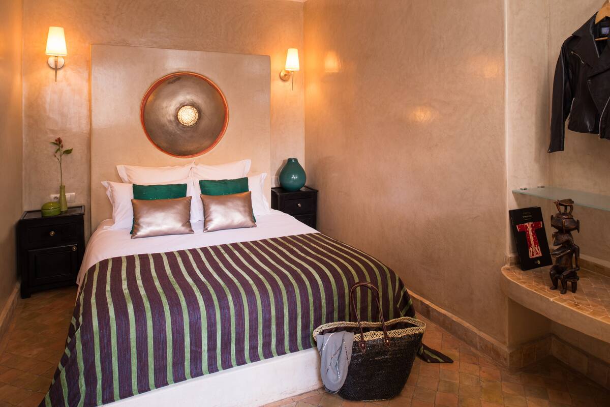A hotel room with a large bed, two bedside tables, a wall-mounted lamp, a vase, a handbag, and a shelf with decorative items.