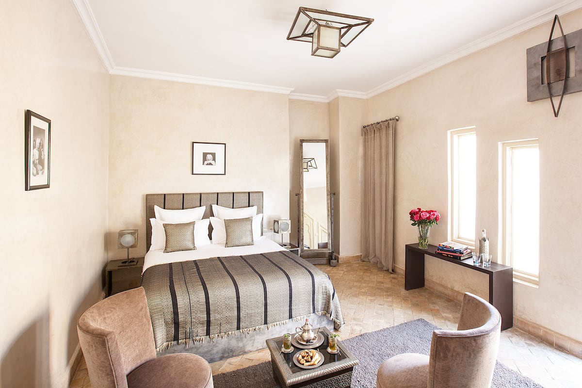 A bedroom with a large bed, a bedside table with a lamp, a chair, a small table with a tray of food, and a window with curtains.