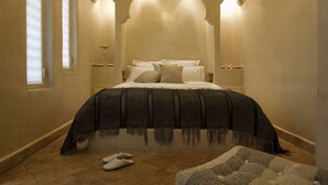 A bedroom with a large bed, two wall-mounted lamps, a window with blinds, and a pair of slippers on the floor. - Riad Cocoon (Marrakech)