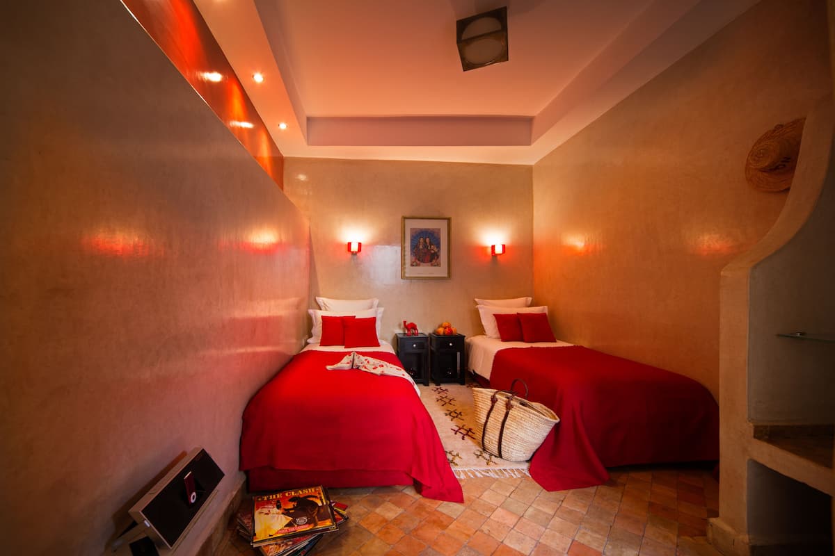 A hotel room with two beds, red bedding, wall-mounted lights, a framed picture, and a small table with a basket.
