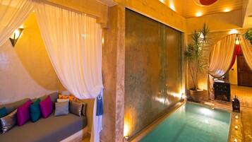 A spacious indoor pool area with a sofa and a small table, surrounded by warm lighting and a large decorative wall.