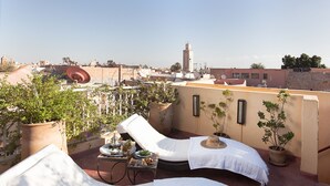 A rooftop terrace with lounge chairs, a table with food and drinks, potted plants, and a view of the cityscape including a tower. - Riad Cocoon (Marrakech)