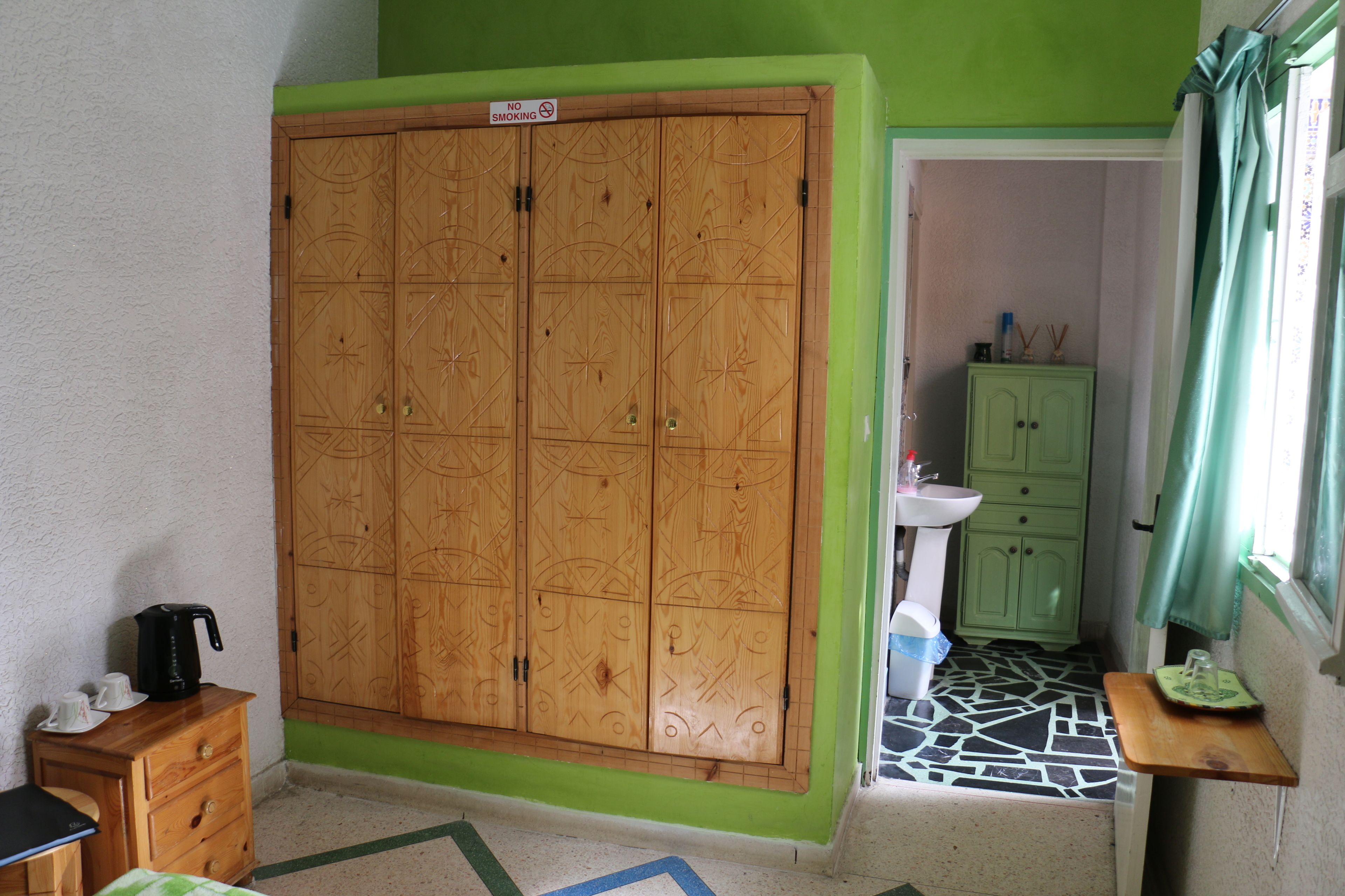 Een kamer met een grote houten garderobe, een klein houten tafeltje met een waterkoker, een groen kastje en een betegelde vloer.