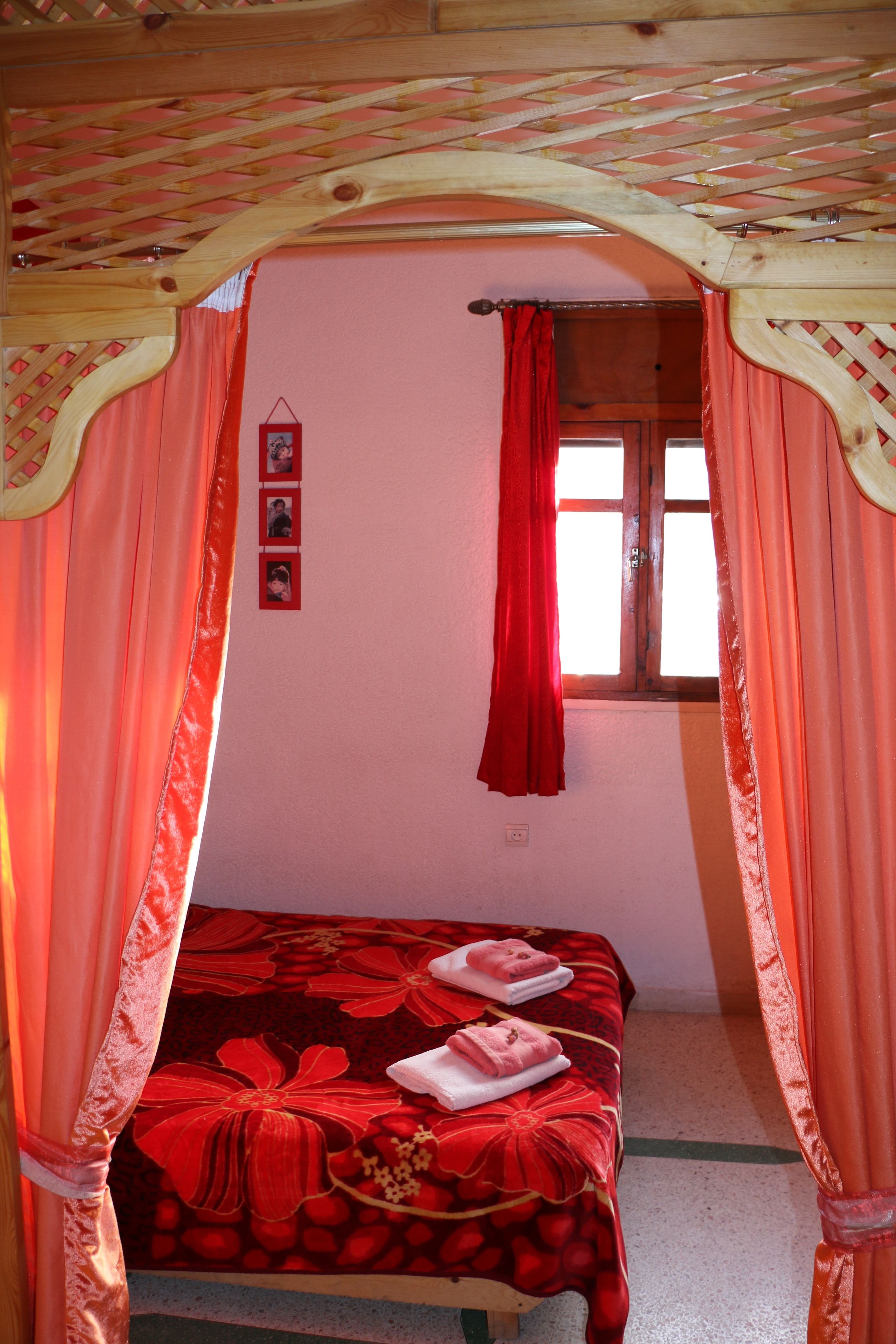 Un dormitorio con cama a dosel, una ventana con cortinas rojas y un muro con tres cuadros enmarcados.
