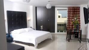 A modern bedroom with a large bed, a dark headboard, a bedside table, a glass coffee table, and a view of the outside balcony. - Hotel Calima Real (Cali)
