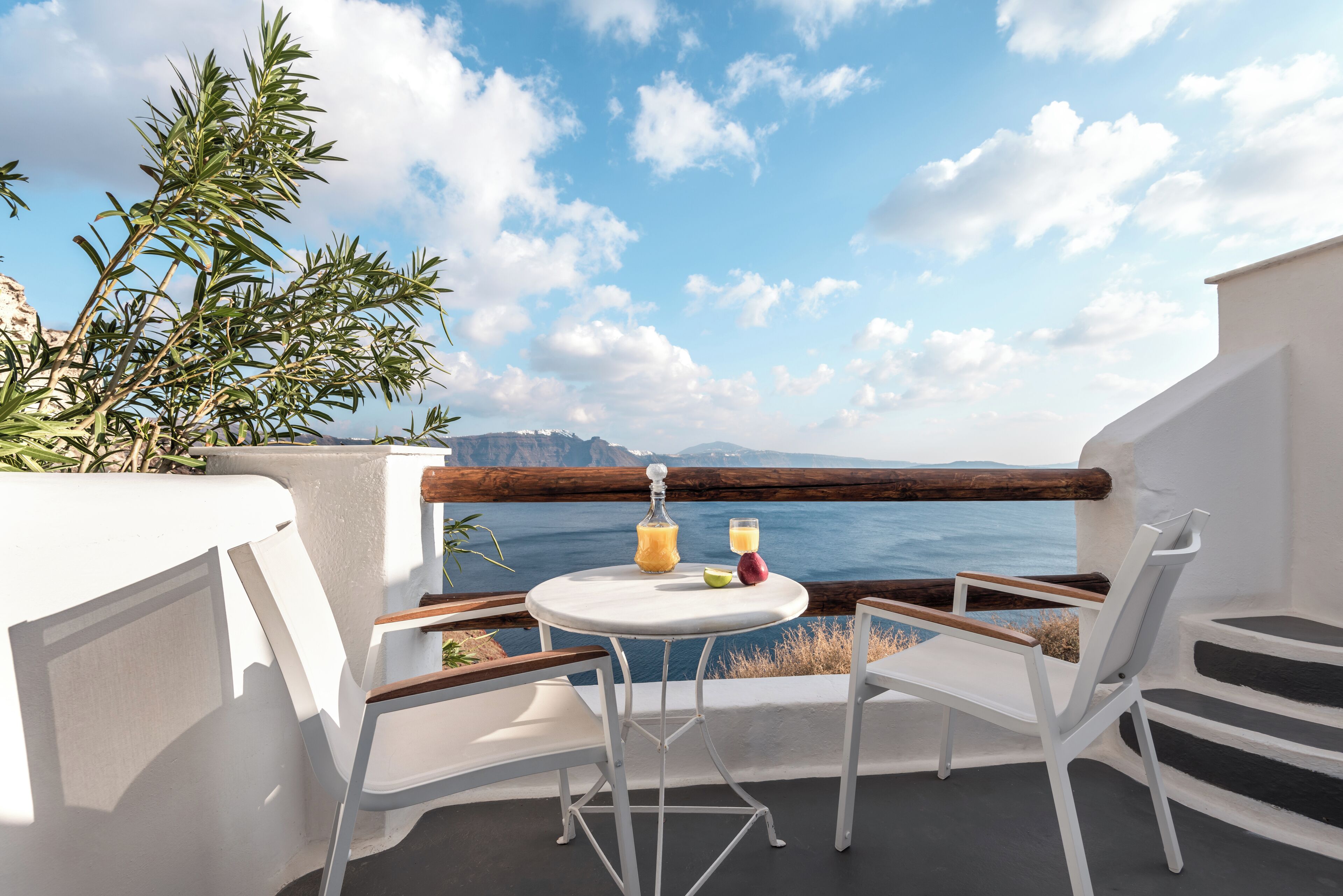 A balcony with white furniture, a table with drinks, and a view of the sea and mountains.