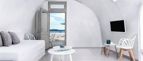 A modern, minimalist room with a white curved ceiling, a sofa with cushions, a small round table with a vase, two chairs, and a TV mounted on the wall.