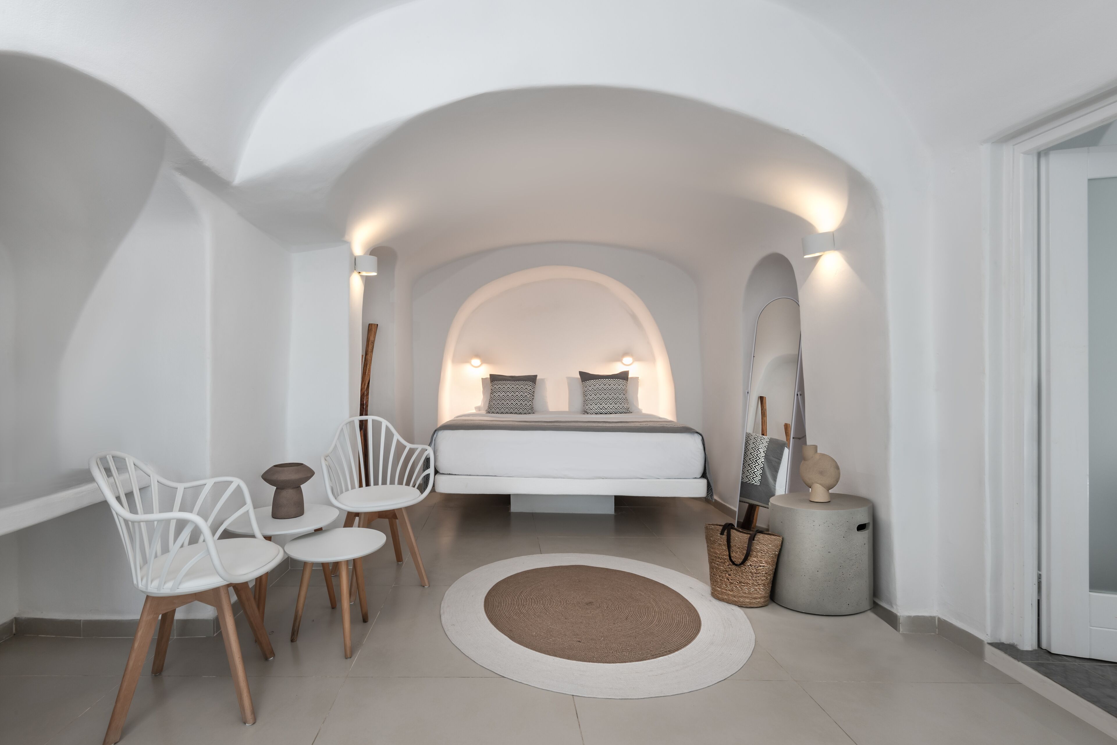 Un dormitorio moderno con una cama centrada bajo un techo blanco en arco, flanqueada por dos lámparas de pared. Se aprecia una alfombra redonda, una mesita redonda con sillas y una cesta decorativa.