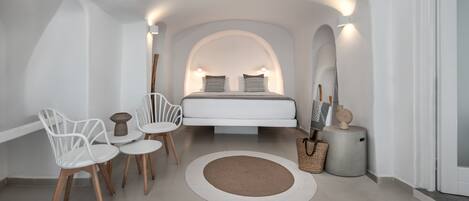 Un dormitorio moderno con una cama centrada bajo un techo blanco en arco, flanqueada por dos lámparas de pared. Se aprecia una alfombra redonda, una mesita redonda con sillas y una cesta decorativa.