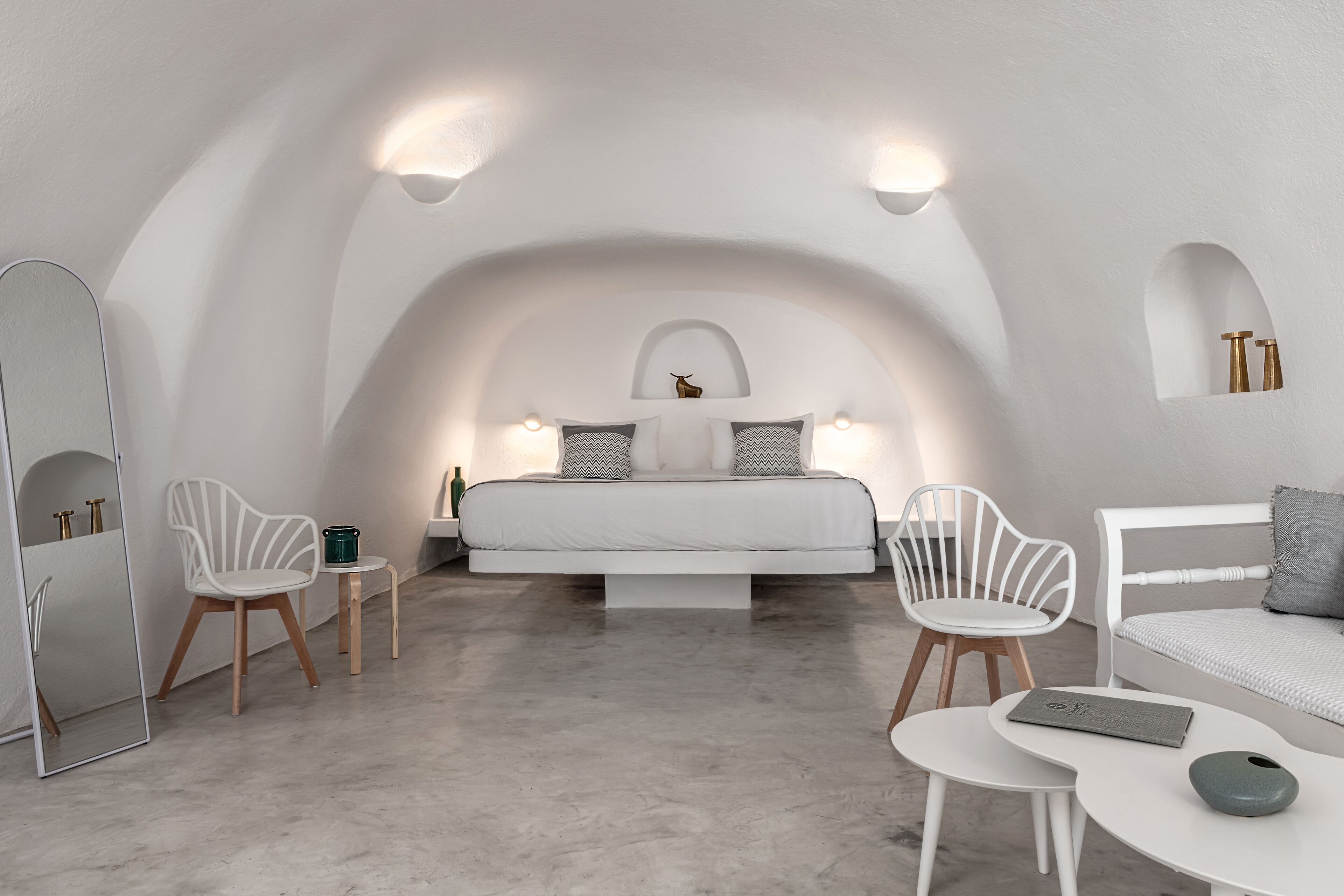 Un dormitorio moderno y minimalista con techo curvo, una cama con ropa de cama blanca, dos sillas blancas, una mesita pequeña y una mesita de noche con lámpara.