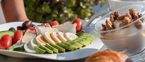 A plate of food with eggs, tomatoes, cucumbers, and cheese, a bowl of yogurt with granola, and a croissant on a table.
