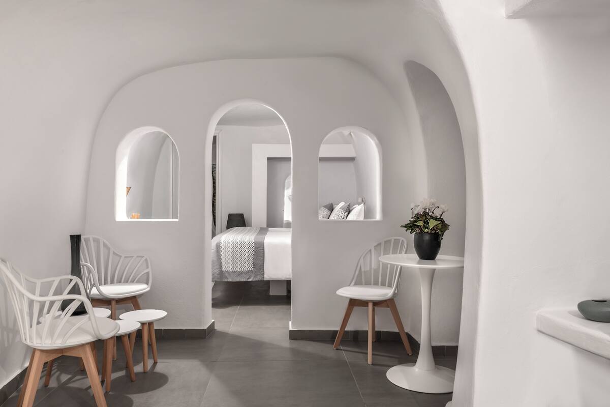A modern, minimalist hotel room with white walls, a gray tiled floor, and a bed with a gray and white patterned bedspread. There are two white chairs with wooden legs and a small white table with a potted plant.