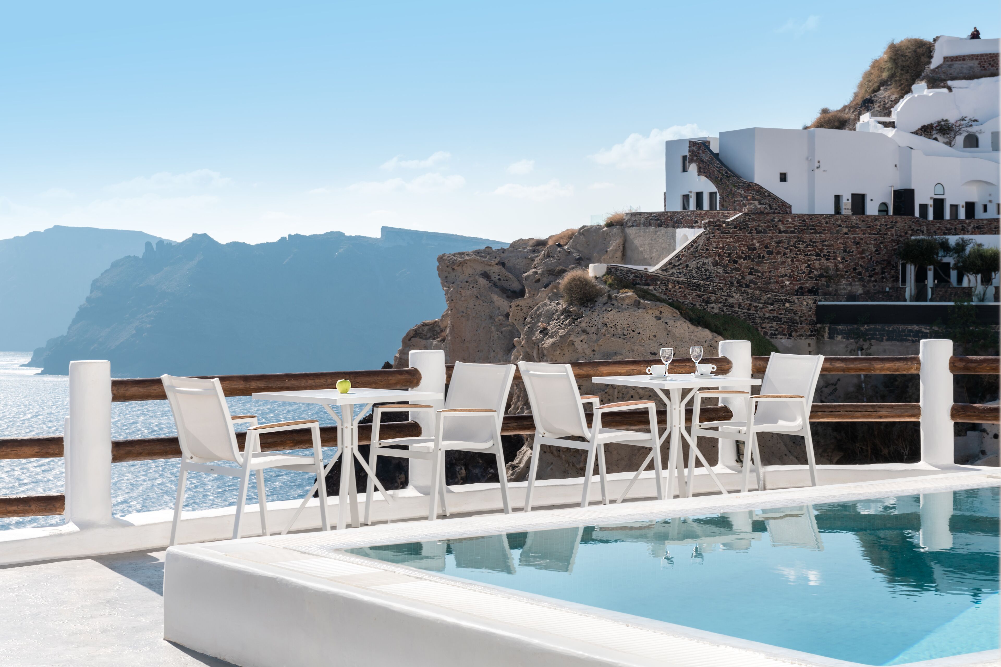 Zona de piscina con sillas y mesas blancas, con vistas a un cuerpo de agua y un edificio blanco con una escalera de piedra.