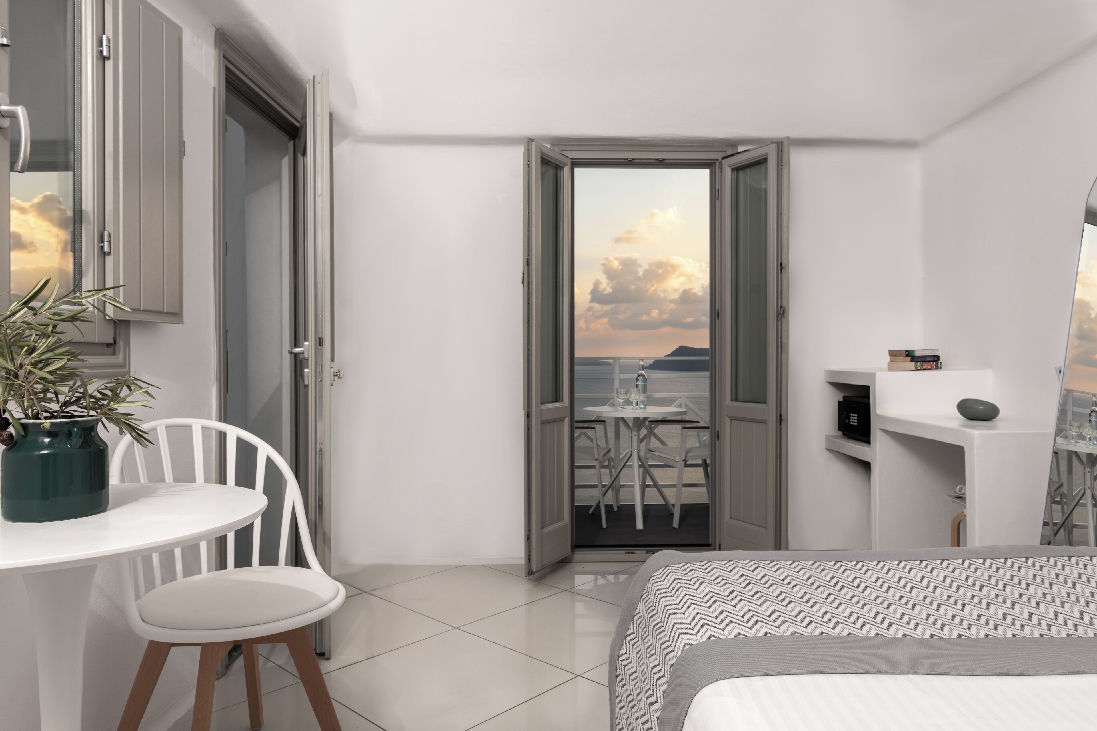 Une chambre moderne avec un lit, une petite table accompagnée d’une chaise, une plante en pot et une vue sur un balcon avec une table et des chaises.