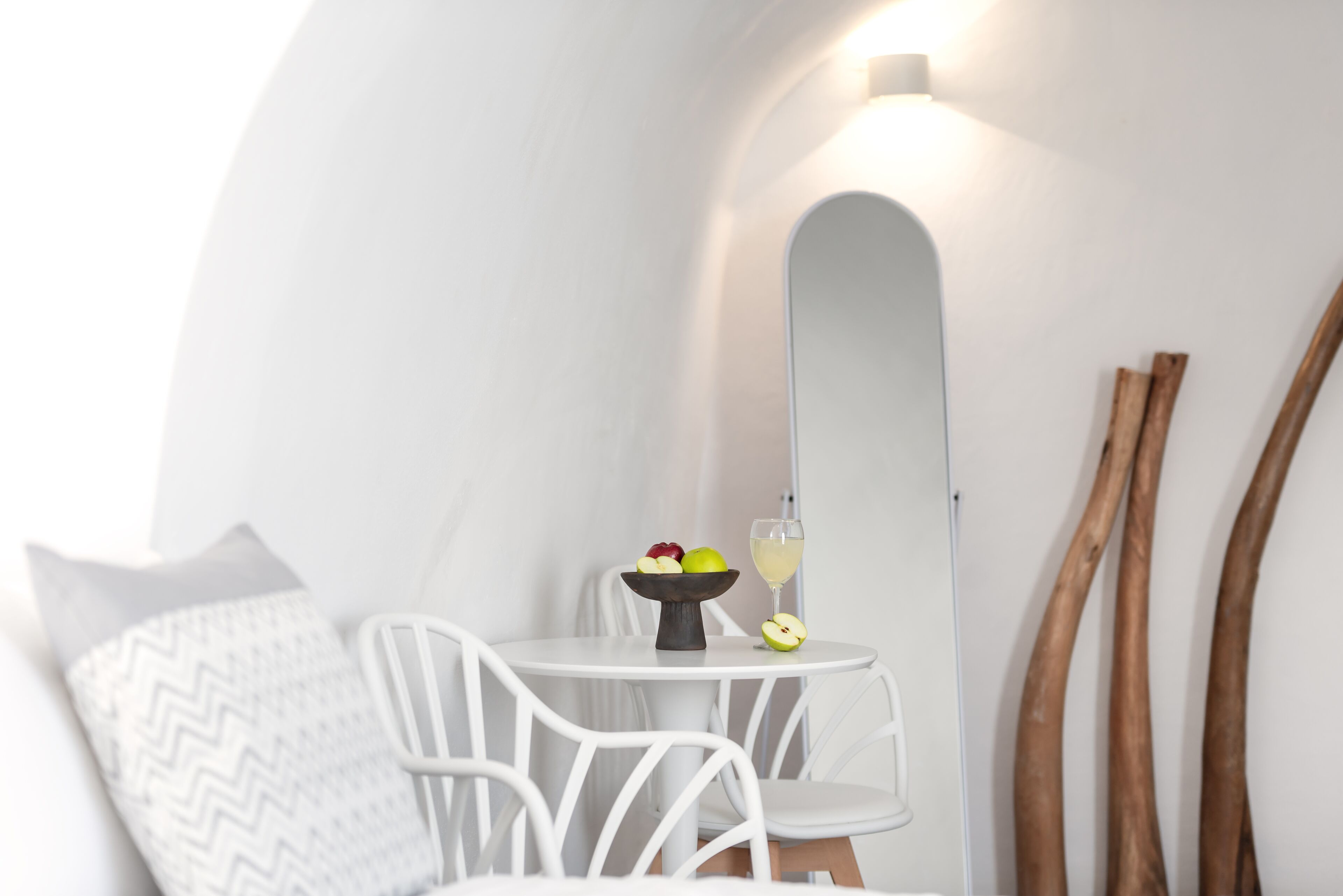 A small, round white table with a bowl of fruit and a glass of drink, a white chair, and a large mirror on the wall.