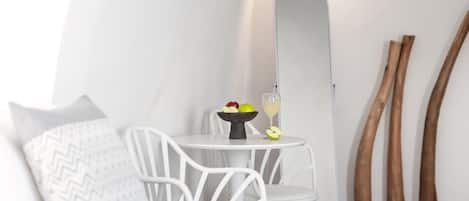 A small, round white table with a bowl of fruit and a glass of drink, a white chair, and a large mirror on the wall.