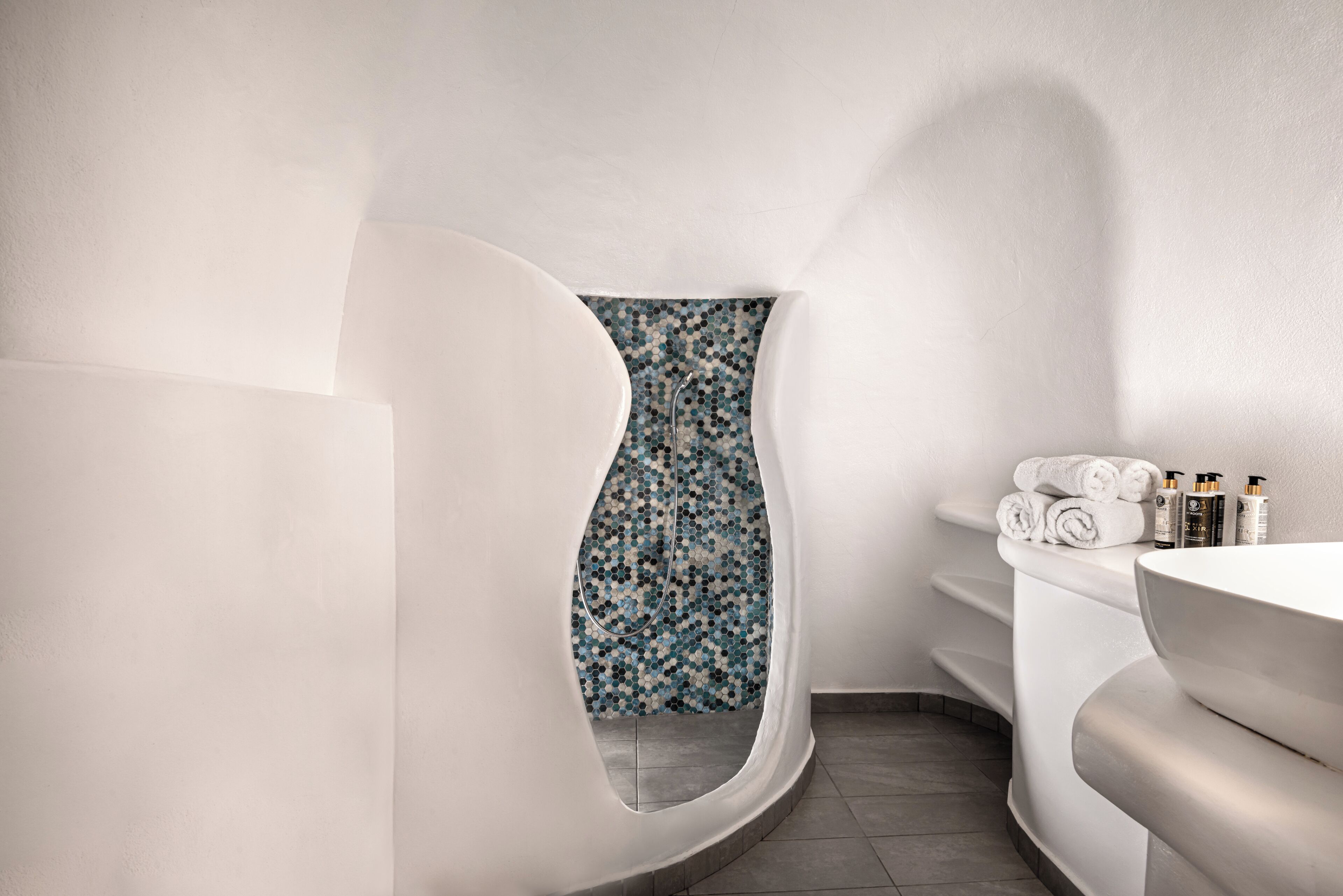 Ett modernt badrum med en unik, böjd design, en duschvägg i mosaik och ett vitt handfat med plats för toalettartiklar.