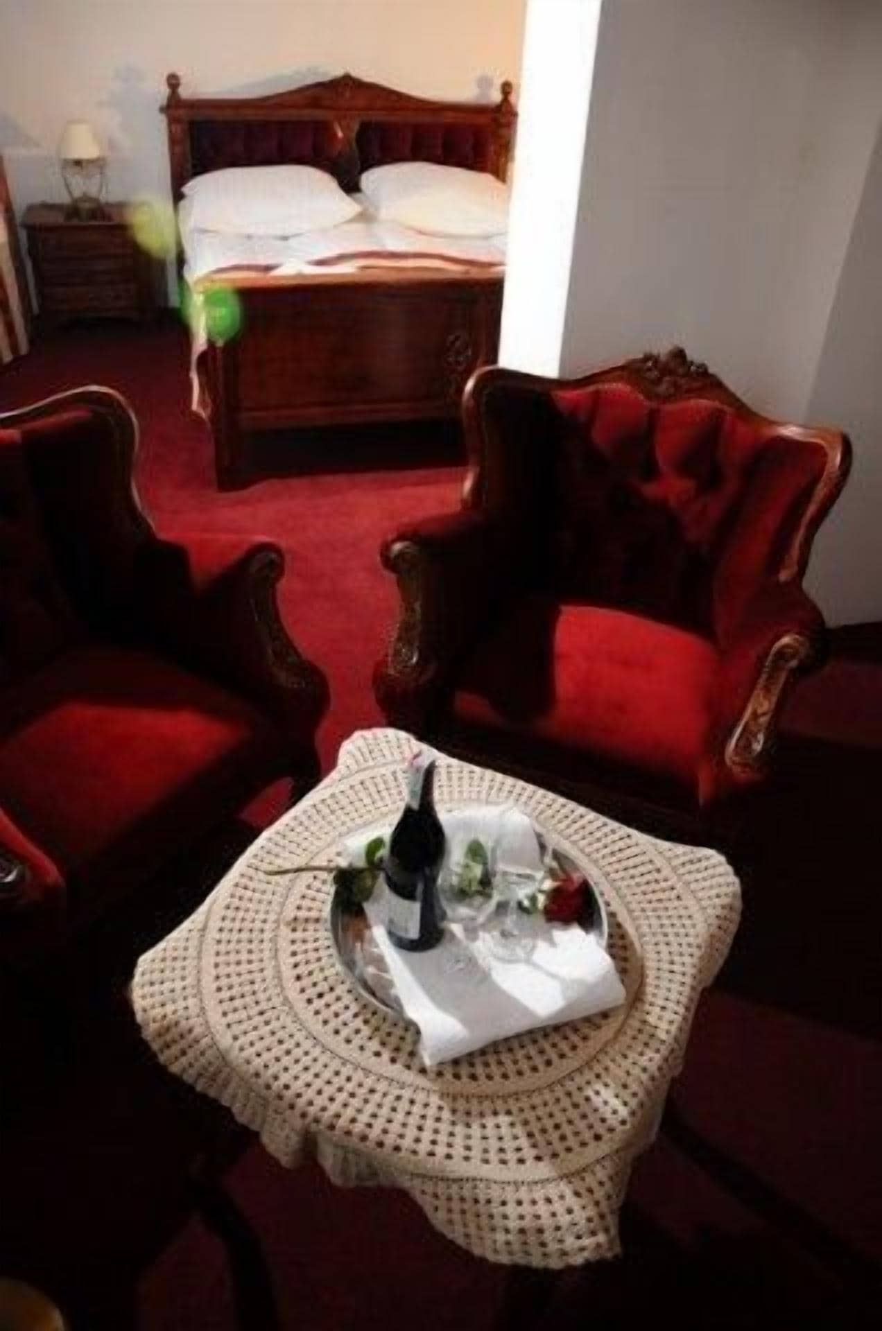 Una habitación de hotel con una cama, dos sillones rojos, una mesita con una botella y vasos, y una alfombra roja.
