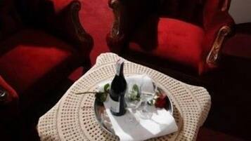 Una habitación de hotel con una cama, dos sillones rojos, una mesita con una botella y vasos, y una alfombra roja.