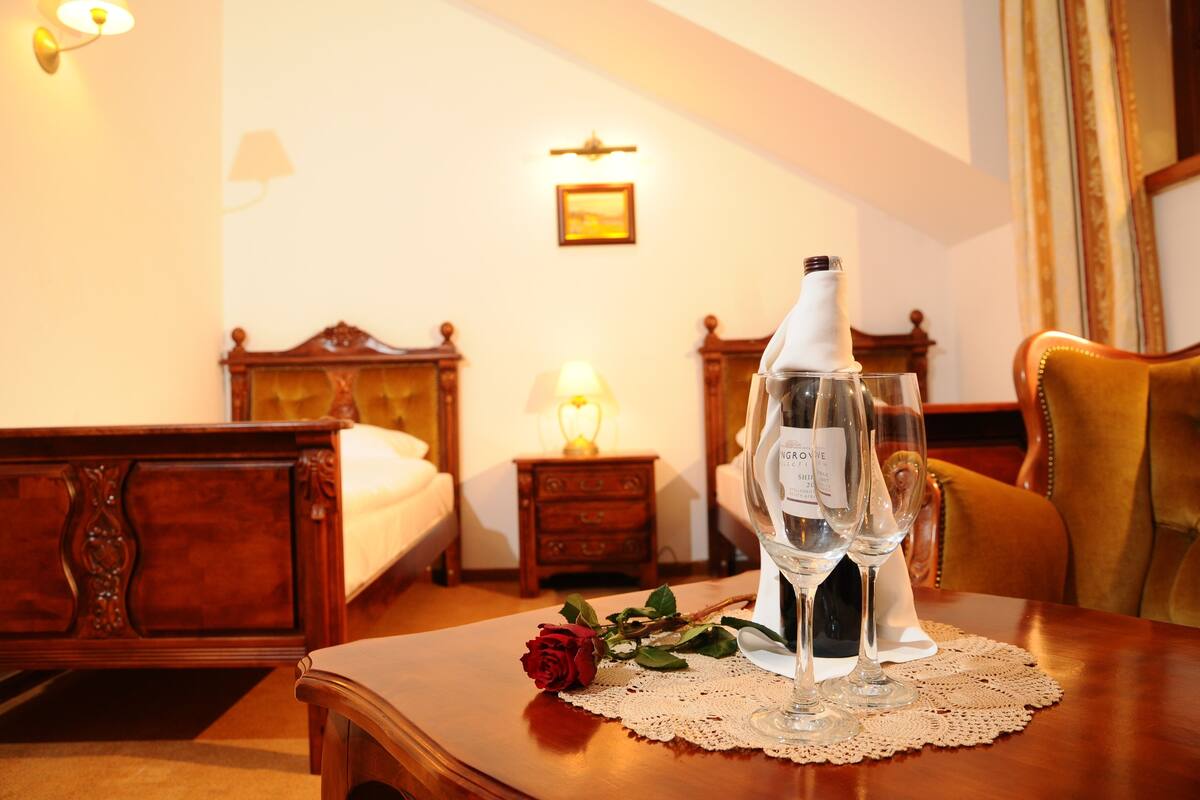 Una habitación de hotel con una cama de madera, una mesita de noche con una lámpara, una mesa con una botella de vino y dos copas, y una rosa roja sobre la mesa.