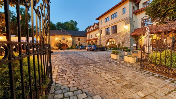 Un camino de adoquinado que conduce a un edificio de piedra con un estilo arquitectónico tradicional, una entrada con portones y coches estacionados.