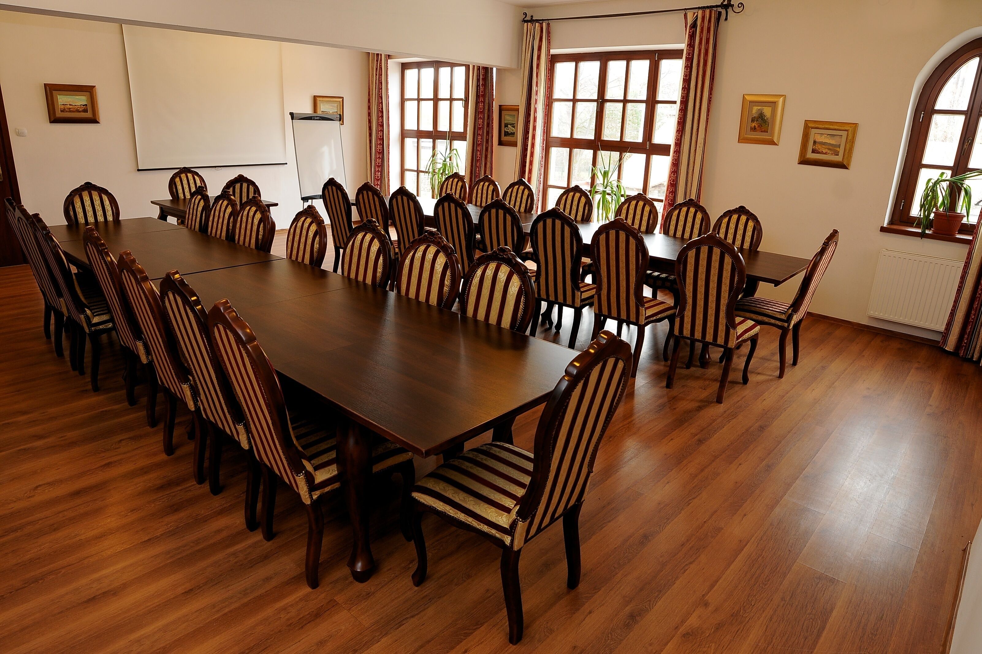 Un salón de conferencias con una larga mesa de madera y sillas a rayas, ventanas amplias con cortinas y cuadros en las paredes.