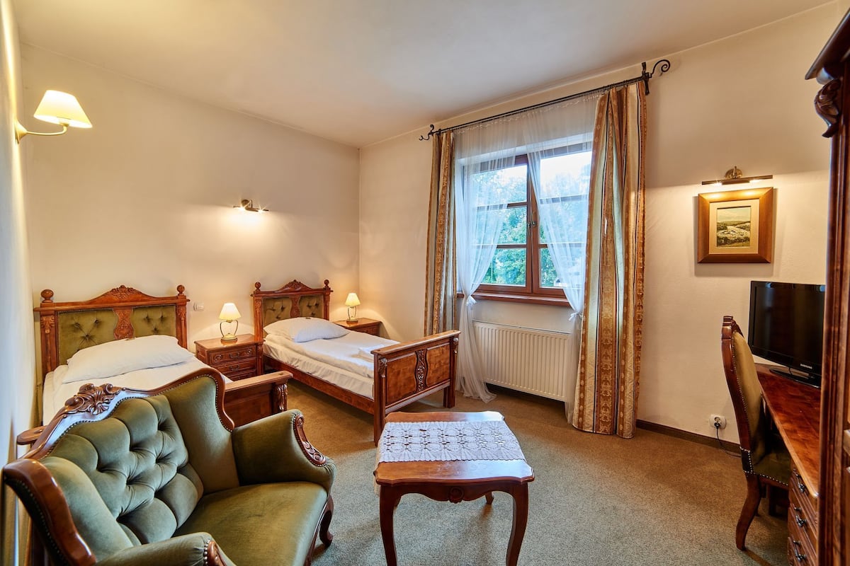 Habitación de hotel con dos camas, un sillón verde, una mesa de madera, un televisor y una ventana con cortinas.